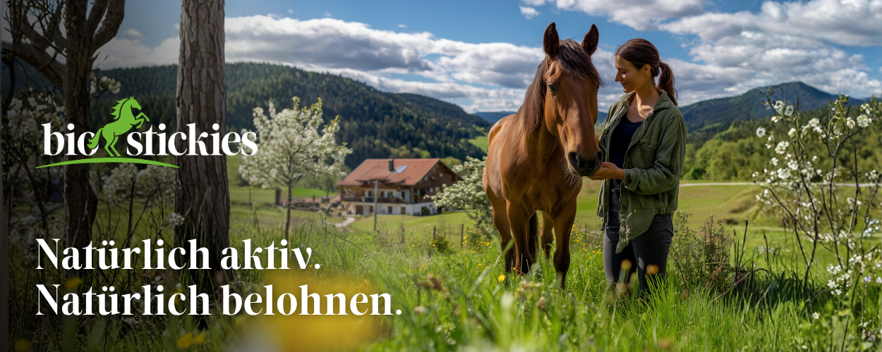 Junge Frau f&uuml;hrt ihr braunes Pferd &uuml;ber eine bl&uuml;hende Fr&uuml;hlingswiese mit Bauernhof und Bergen im Hintergrund &ndash; biostickies: Nat&uuml;rlich aktiv. Nat&uuml;rlich belohnen.
